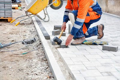 A bricklayer in a blue and orange overalls, half-squatting, taps the paving slabs.