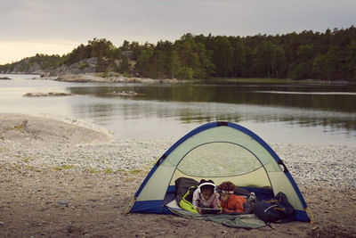 Siblings with digital tablet lying in tent at beach during sunset