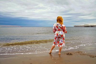 Rear view of woman standing at beach against cloudy sky