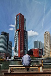 Rear view of man sitting on bench by river against buildings