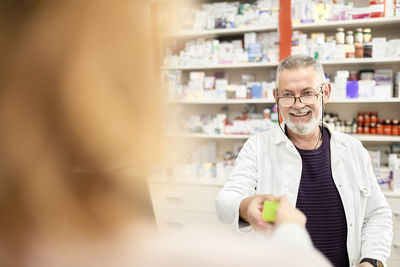 Smiling pharmacist wearing eyeglasses giving medicine to customer at pharmacy store