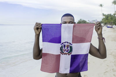 A young black boy stands at the edge of the caribbean sea,holding the flag of the dominican republic