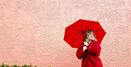 Woman looking away while holding red umbrella against wall