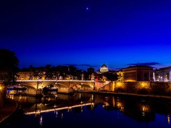 Illuminated bridge over river by buildings against sky at night