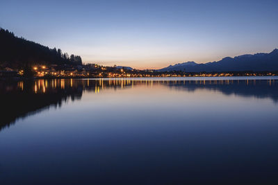 Illuminated town reflecting in lake hopfen against clear sky at dusk