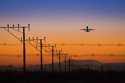 Poles on field against silhouette airplane flying in sky during sunset