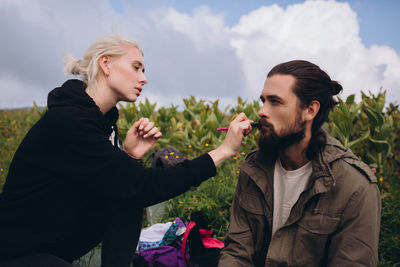 Woman applying make-up on man while sitting outdoors