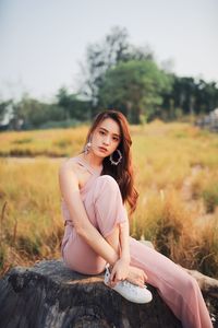 Portrait of young woman sitting on tree stump against clear sky