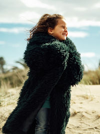 Side view of smiling little girl wrapped in fur coat with eyes closed standing in wind on sandy beach in sunny day