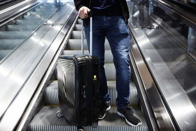 Low section of man with wheeled suitcase standing on escalator