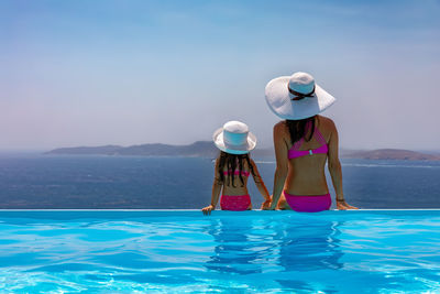 Rear view of mother and daughter looking at sea while sitting by swimming pool against sky