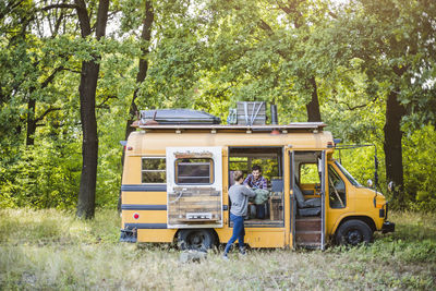 Woman helping man in unloading luggage from yellow caravan during camping in forest