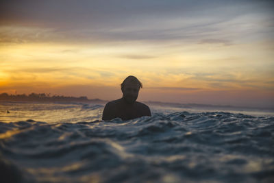 Man sitting on surfboard in sea during sunset