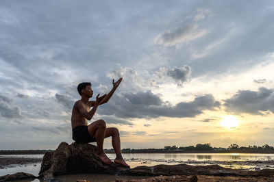Shirtless boy sitting on rock against sky during sunset