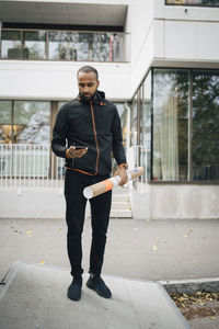 Thoughtful male worker with package using smart phone while standing against building in city