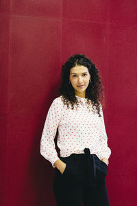 Portrait of confident businesswoman with hands in pockets standing by red wall in creative office