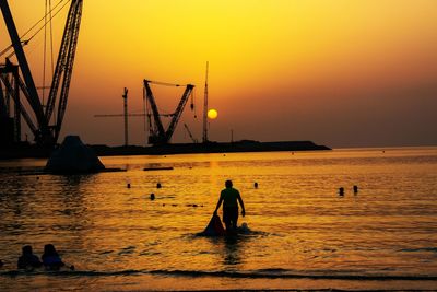 Silhouette men sitting on sea against orange sky