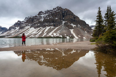 Rear view of woman standing at lakeshore against mountains during winter