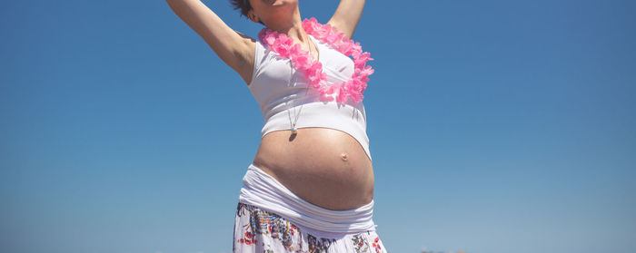 Midsection of pregnant woman with arms raised against clear sky