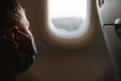 Kid in face mask admiring view behind plane window while travelling during epidemic