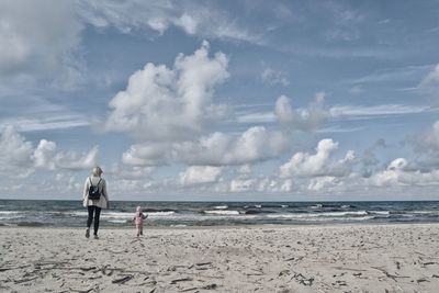 Rear view of mother and daughter walking on beach against sky