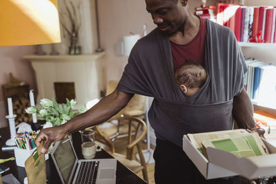 Male professional packing box while carrying baby boy in carrier at home office