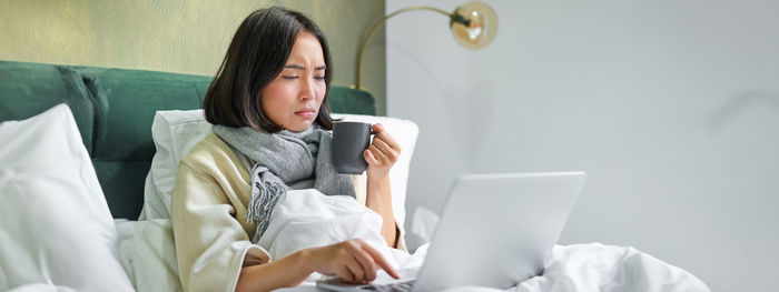 Young woman using digital tablet while sitting on bed at home