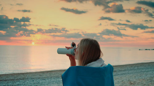 Rear view of woman drinking coffee at beach during sunset