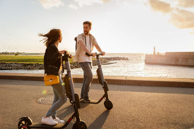 Couple talking while riding electric push scooters on road by sea against sky