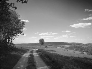 Dirt road along trees on field against sky