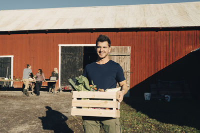 Portrait of smiling mid adult man carrying crate full of organic vegetables with barn in background