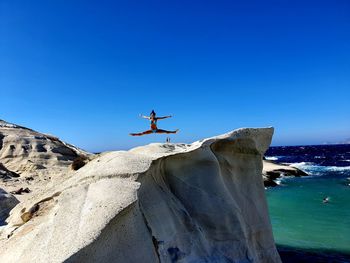 Young girl jumping on rock by sea against clear blue sky