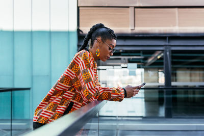 Side view of young ethnic female in stylish colorful clothes leaning on glass fence and browsing social media on smartphone on terrace outside modern building