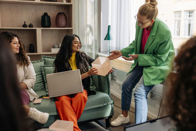 Smiling businesswoman showing box to colleague while sitting with laptop at office