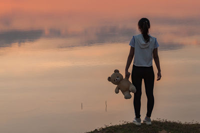 Rear view of woman holding teddy bear while standing against lake during sunset