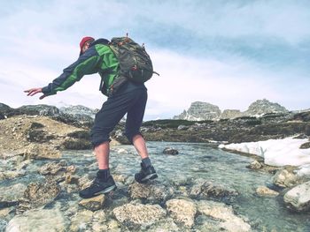 Man with backpack and in trekking boots is crossing carefully over mountain river. snowy mountains