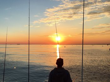 Rear view of man fishing while standing by sea against sky during sunset