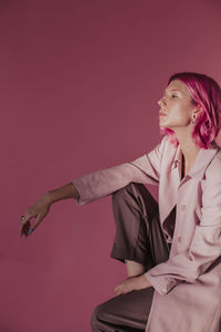Young woman looking away while sitting against pink background