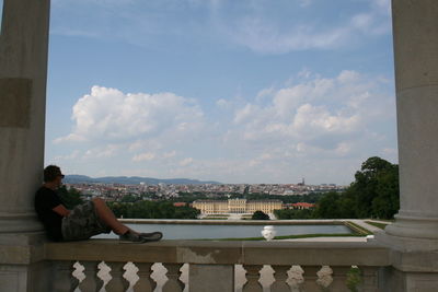 Man sitting on bridge over cityscape against sky