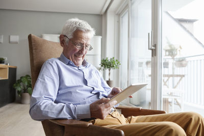Portrait of happy senior man sitting on armchair at home using digital tablet
