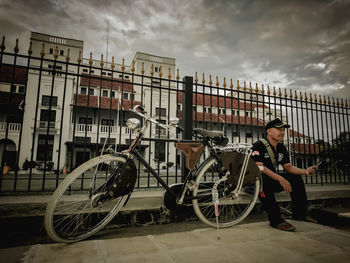 Man with bicycle standing by railing in city against sky