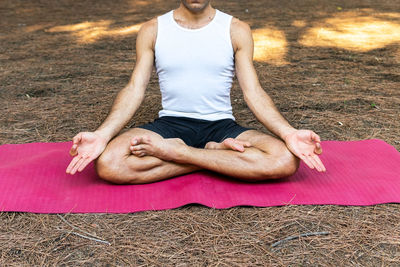 Unrecognizable barefoot male sitting in lotus pose on magenta mat and gesturing mudra during meditation session on sunny summer day in park