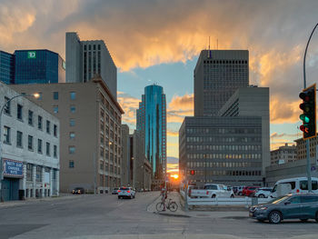 Cars on city street by buildings against sky during sunset