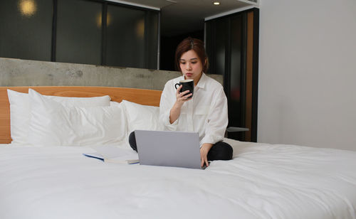 Full length of woman holding coffee cup using laptop sitting on bead at home