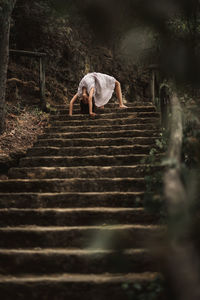 Young female in white dress standing in weird bridge pose leaning on hands on stairs doing yoga exercise in green park