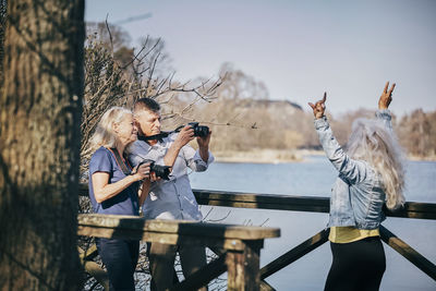 Senior man and woman photographing friend through camera while standing by lake