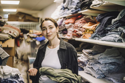 Portrait of young female fashion designer standing near clothes shelf at workshop