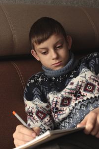 A boy in a christmas sweater sits on a sofa and draws with a pencil in his free time