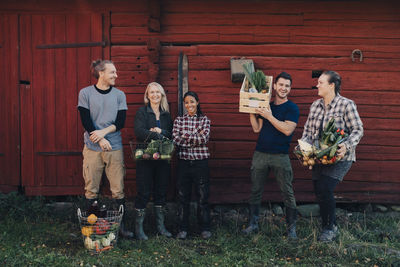 Full length of multi-ethnic farmers with organic vegetables standing against barn