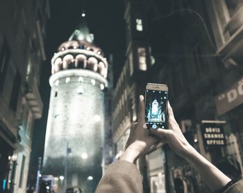 Cropped hands of woman photographing galata tower in city at night